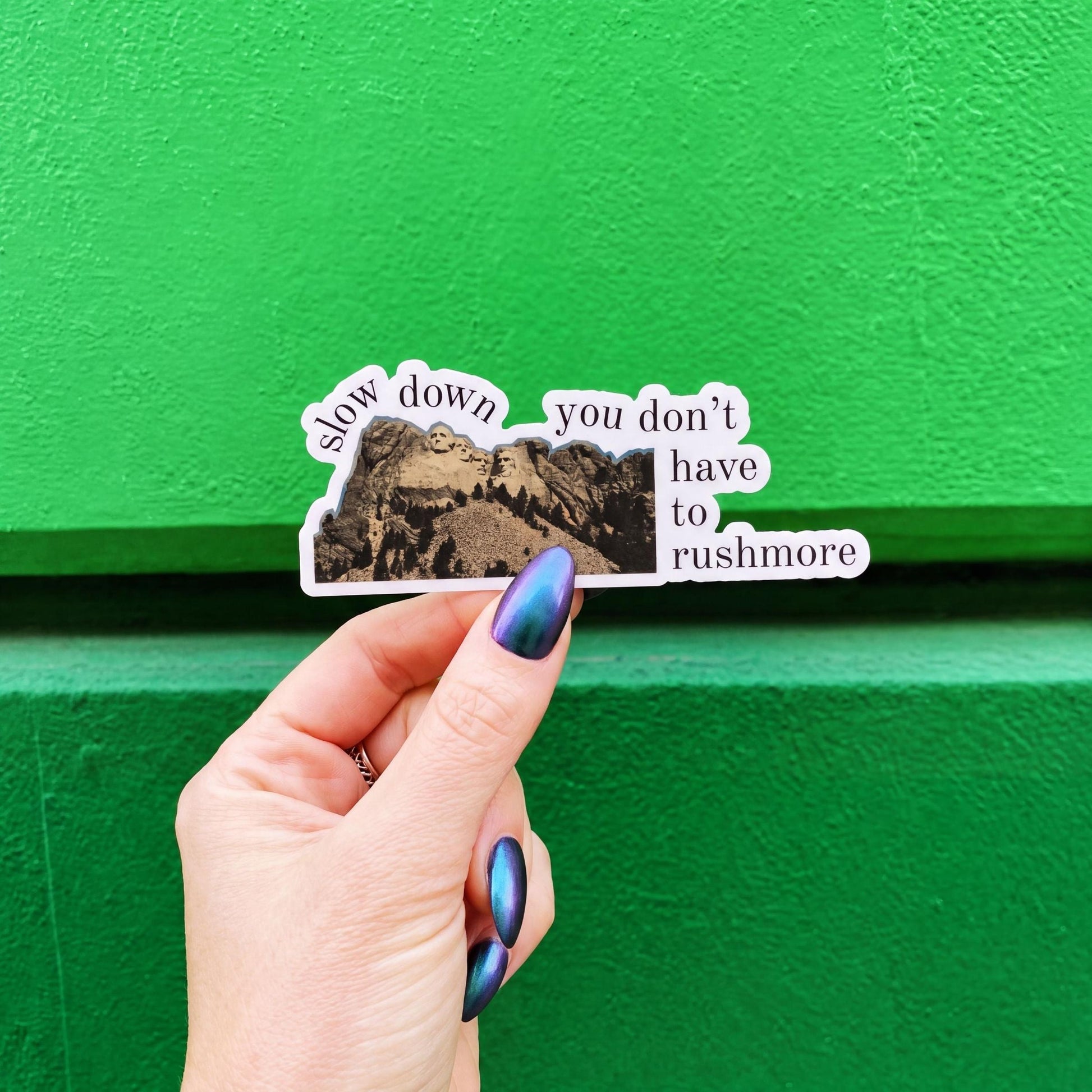 A die-cut sticker featuring Mount Rushmore and the text 'slow down you don't have to rushmore' 
